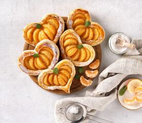 Puff pastry cake in the shape of a heart with tangerine slices on a wooden board on a white table with napkin. top view, copy space