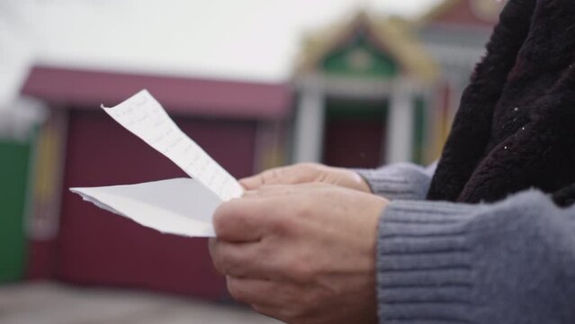 Hands of elderly man holding letter and envelope from friend on blurred background. Pensioner stands in yard on snowy winter day closeup