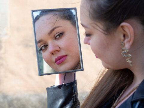 Mujer joven posando, mir&aacute;ndose al espejo en el exterior, con la cara blanca, mirada fija, labios rojos, en primavera de 2021