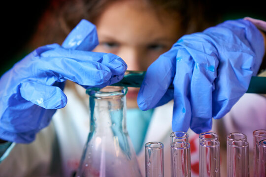 Little kid doing experiments with blue liquid. Cute child wants to become a doctor. Pretty child in white medical robe holds glass flasks with blue liquid on indigo background with hospital symbol. - Powered by Adobe