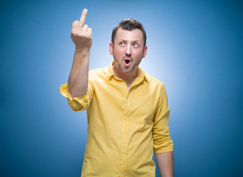 Young man showing fuck off gesture over blue background, dresses in yellow shirt. Male person with obscene gesture giving the middle finger and looking to camera