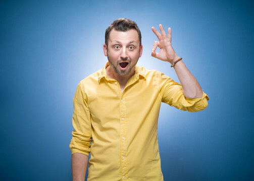 Surprised young man shows excellent sign with finger over blue background, dresses in yellow shirt. Yes or excellent gesturing, body language concept