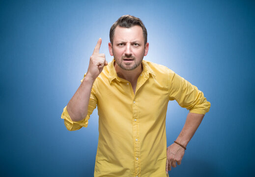 Angry man pointing finger up with the index finger a showing problem over blue background, dresses in yellow shirt. Fury guy screaming