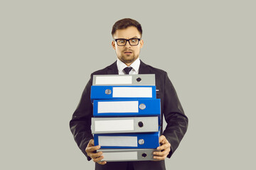 Bureaucracy and overworking concept. Angry office worker with pile of papers and documents is stressed by lot of work and deadlines. Man in suit angrily looks at stack of folders on gray background.