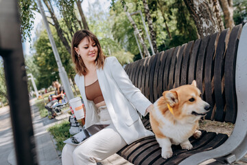 Pretty businesswoman in white suit sitting in city parkland, drink coffee and working on digital tablet with her dog Welsh Corgi Pembroke