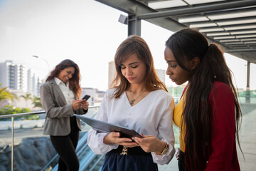 Two female colleagues work using a tablet while the other colleague in the background uses her smartphone.