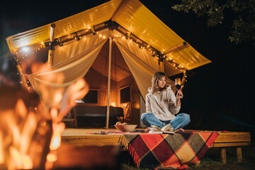 Fototapeta na wymiar Smiling Woman freelancer drinking wine and read book sitting in cozy glamping tent in autumn evening. Luxury camping tent for outdoor holiday and vacation. Lifestyle concept