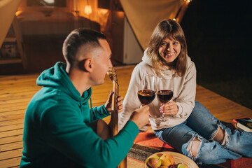 Happy couple relaxing in glamping on autumn evening, drinking wine and playing guitar near cozy bonfire. Luxury camping tent for outdoor recreation and recreation. Lifestyle concept