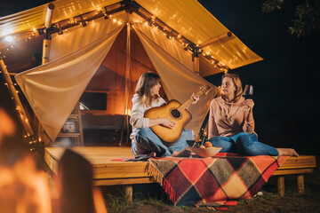 Obraz na płótnie Canvas Two Smiling female friends drinking wine and play guitar sitting in cozy glamping tent in autumn evening bonfire. Luxury camping tent for outdoor holiday and vacation. Lifestyle concept