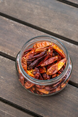 A clear glass jar without a lid full of dried chilli peppers on acacia wood surface