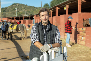 Portrait of middle-aged male stable worker smiling at camera during work in horse club on sunny...