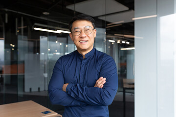 Portrait of successful asian boss inside office, businessman with crossed arms smiling and looking at camera, man in shirt and glasses happy with result of achievement at work.
