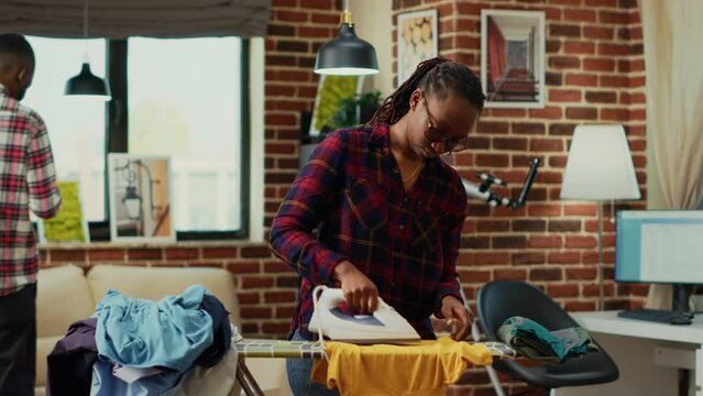 Young modern housewife using heat iron and ironing table, smoothing out creases on clothes. Female person doing household chores for spring cleaning, using laundered clothing.