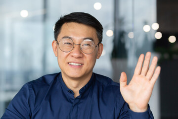 A successful Asian man inside the office is looking at the web camera, talking on a video call with colleagues, gesturing and smiling an online meeting with partners, talking to customers, pov