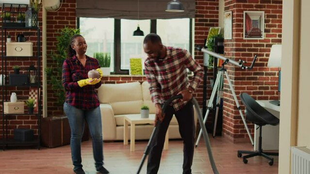 Young life partners doing household chores and cleaning apartment with vacuum cleaner. African american woman sweeping dust off of furniture with rags and washing solution, furniture polish.