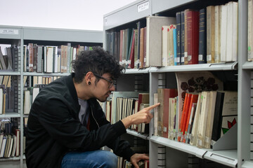 Estudiante latino de cabello crespo, gafas y expansiones buscando un libro en la biblioteca