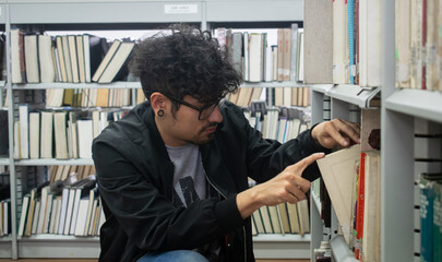 Estudiante latino de cabello crespo, gafas y expansiones buscando un libro en la biblioteca
