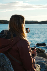 Fototapeta na wymiar Blonde girl drinks hot tea on the beach. Young hiker takes a coffee break with a thermal flask. Moment of reflection and peace of mind. Comfort, peace and hot drinks.