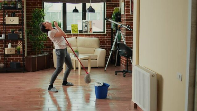 Caucasian wife cleaning apartment floors with mop, listening to music and doing chores. Cheerful woman dancing and washing dirt, doing housework in living room. Spring cleaning.