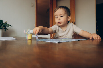 child draws with a brush and multicolored gouache sitting at the table drawing to his mother