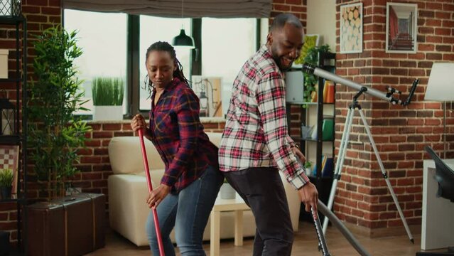 Cheerful people showing dance moves and doing spring cleaning, having fun with music while they clean wooden floors with mop and vacuum cleaner. Young couple smiling and doing chores.