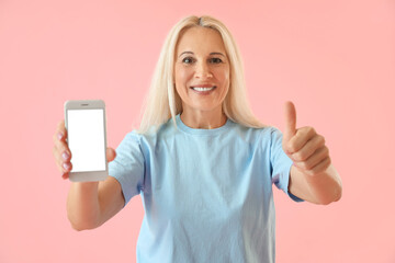 Mature blonde woman with mobile phone showing thumb-up on pink background