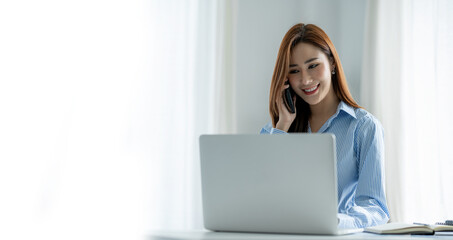 Happy young Asian woman talking on the mobile phone and smiling while sitting at her working place in the office.