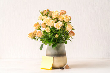 Vase with beautiful rose flowers and blank card on table near light wall