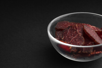 Bowl with pieces of delicious beef jerky on black table, closeup. Space for text