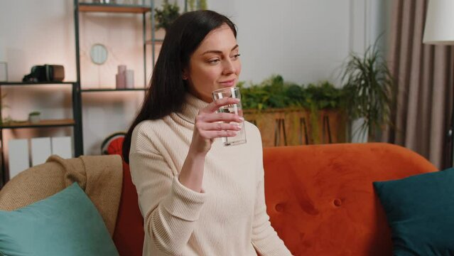 Thirsty brunette woman holding glass of natural aqua make sips drinking still water preventing dehydration sits at home living room. Girl with good life habits, healthy slimming, weight loss concept