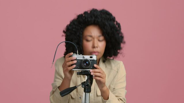 Serious female photographer capturing pictures with on film camera mount on tripod on pink background. Young woman of color using retro camera at studio. Girl making shot on vintage film camera 4K
