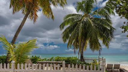 Fototapeta na wymiar The relaxation area on the tropical beach is surrounded by a white fence. A wooden table, benches, and deck chairs are visible. Palm trees against a blue sky, clouds, turquoise ocean. Seychelles