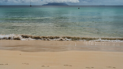 A wave of turquoise ocean foams on the shore, mixing with sand.  Footprints in wet sand. Outlines of the island and yachts on the horizon. Clouds in the blue sky. Seychelles. Mahe. Beau Vallon.
