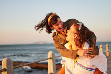 Women, gay couple and piggyback by the ocean coast with smile for happy relationship and fun pride in the outdoors. Lesbian woman on back ride with partner laughing in joy and happiness by the beach - obrazy, fototapety, plakaty