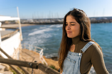 Fototapeta na wymiar Hispanic female with long hair closing eyes delightfully while standing on pier near rippling sea and enjoying sunshine