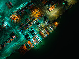night scene shot commercial port loading and unloading cargo from container ship import and export by crane for distributing goods by trailers transported aerial top view