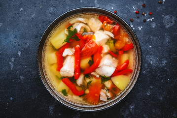 Fish soup with cod, paprika, potatoes, tomatoes and parsley in ceramic soup bowl on dark table background, top view