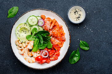 Ketogenic diet salad bowl with salmon, shrimp, avocado, spinach, cucumber, tomato, cashew nuts, sesame. Low-carbohydrate breakfast rich in healthy fats. Black table background, top view