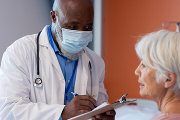 Diverse senior male doctor in face mask talking with senior female patient, making notes, copy space