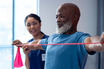 Smiling diverse female physiotherapist helping senior male patient exercise with band