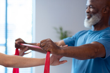 Diverse female physiotherapist helping senior male patient exercise with band, copy space