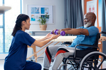 Diverse female physiotherapist helping senior male patient in wheelchair exercise with dumbbells - Powered by Adobe