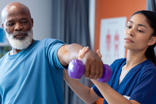 Diverse female physiotherapist helping senior male patient exercise with dumbbell, copy space