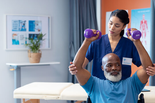 Diverse female physiotherapist helping senior male patient exercise with dumbbells, copy space