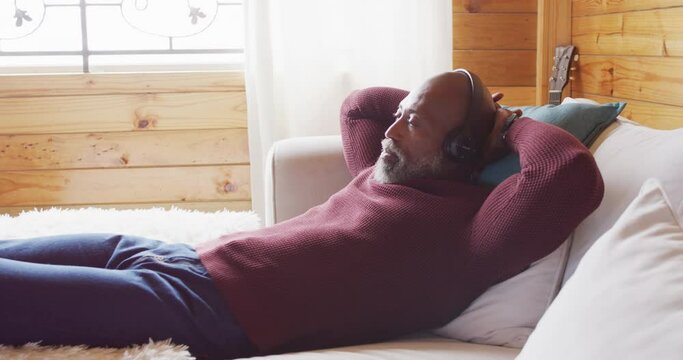 Happy senior african american man in log cabin, laying on sofa and using headphones, slow motion