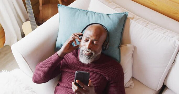 Happy senior african american man in log cabin, laying on sofa and using smartphone, slow motion