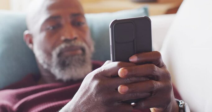Happy senior african american man in log cabin, sitting on sofa and using smartphone, slow motion