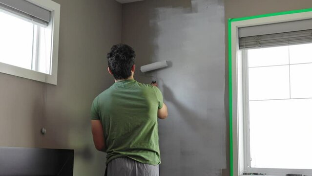 A Caucasian man walks into frame with a paint roller covered in white paint and begins to paint the wall beside a window.