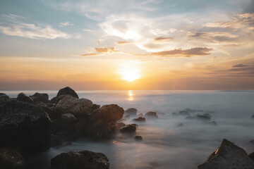 Obraz premium (Long exposure) Stunning seascape with some rocks in the foreground and the sun setting in the distance. Lombok Island, West Nusa Tenggara, Indonesia.