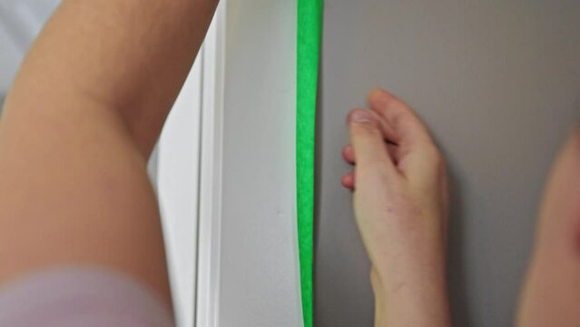 Close up of a white woman applying green painters tape along the edge of a door frame.